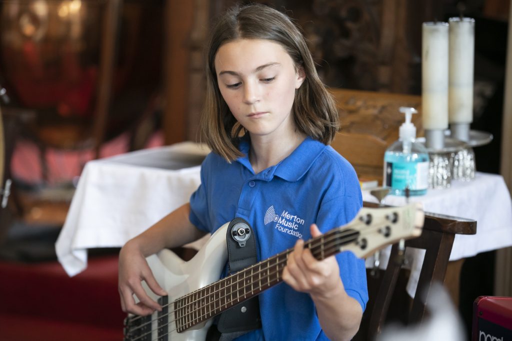 A young person in a blue 'Merton Music Foundation' polo shirt sat down playing a white bass guitar. They are deep in concentration. 