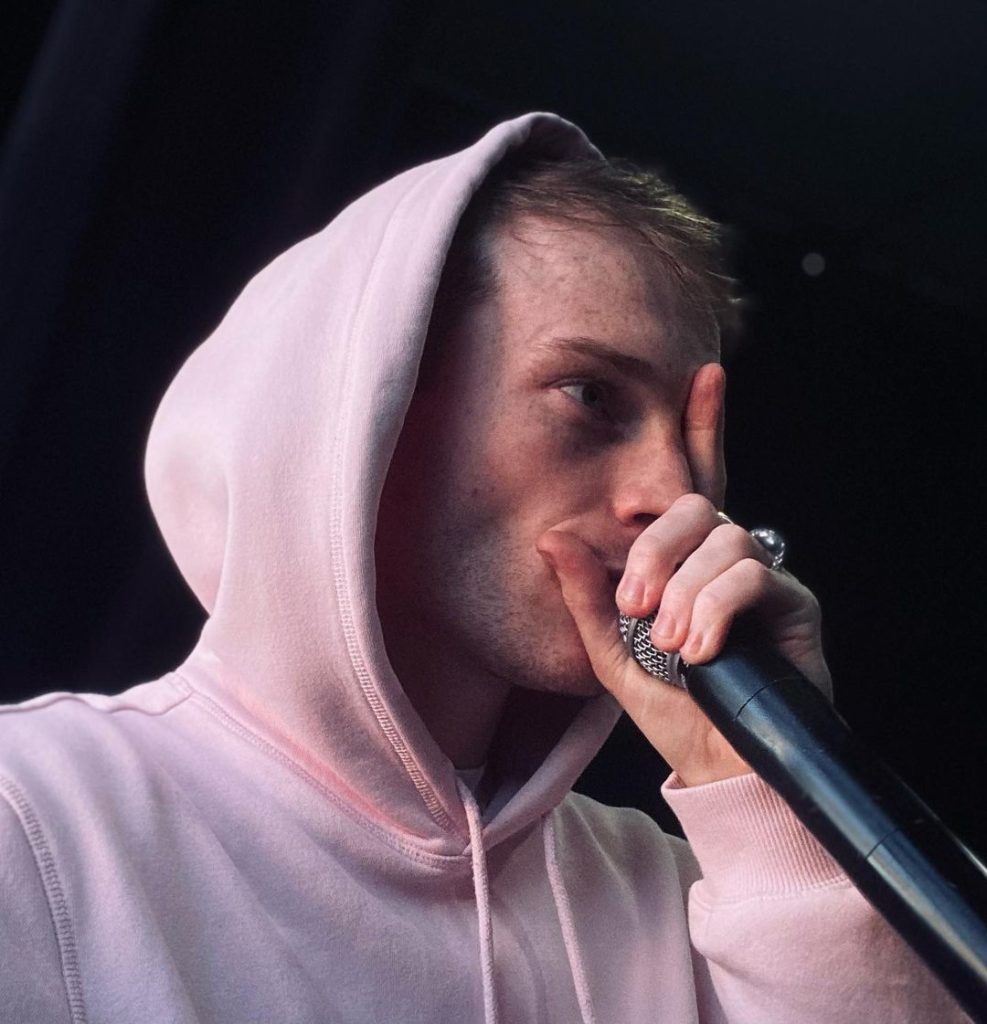 A man in a pink hoodie beatboxes into a microphone. 