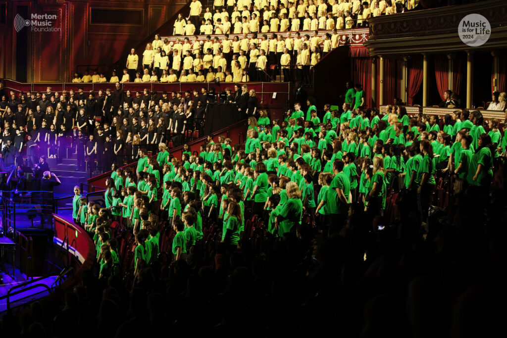 A large choir of children in bright green shirts sings on stage at a concert hall, with other groups in black and yellow attire seated behind them and an audience watching. Banners read Merton Music Foundation and Music is for Life.