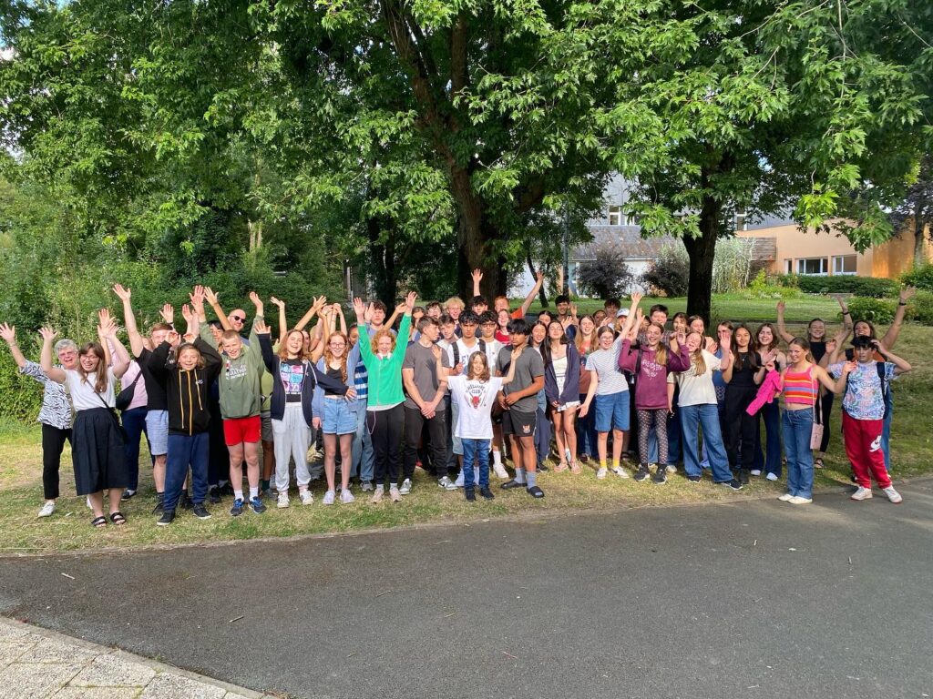A large group of people, mostly young, stand outdoors under trees, smiling and waving enthusiastically at the camera. It’s a sunny day, and the group appears happy and cheerful.