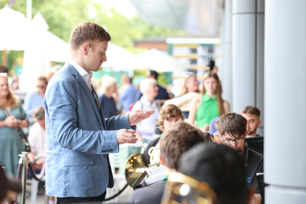 A man in a blue blazer conducts a small band outdoors, with musicians and musical instruments visible; a crowd watches in the background.