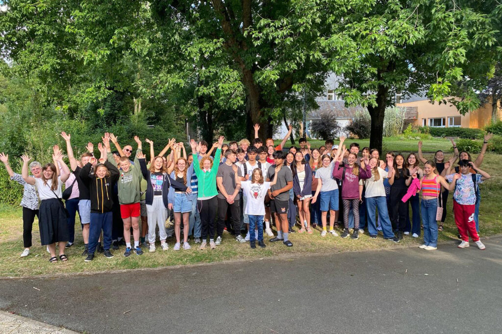 A large group of people, mostly teenagers, stand outdoors on a sunny day, smiling and waving their hands in the air, surrounded by trees and greenery. Some buildings are visible in the background.