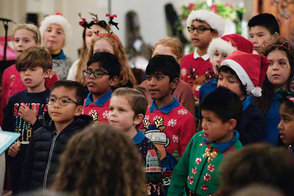 A group of children, some wearing festive jumpers and Santa hats, sing together at a holiday event. The background is decorated with flowers and holiday decorations.