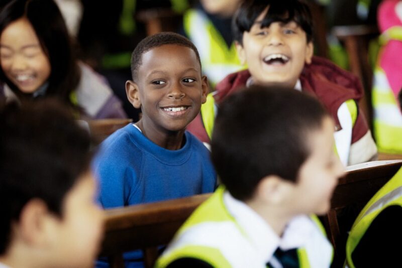 A group of children sit together, smiling and laughing. One boy in a blue shirt is centred and looking at the camera, while others around him appear happy and engaged—a lively, joyful moment perfect for Photo Collections.