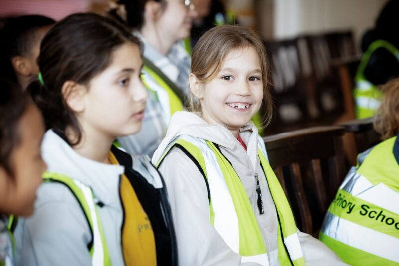 Two young girls sit indoors wearing yellow safety vests over their clothes. One girl smiles at the camera whilst others sit nearby. The atmosphere suggests a school event or group outing.