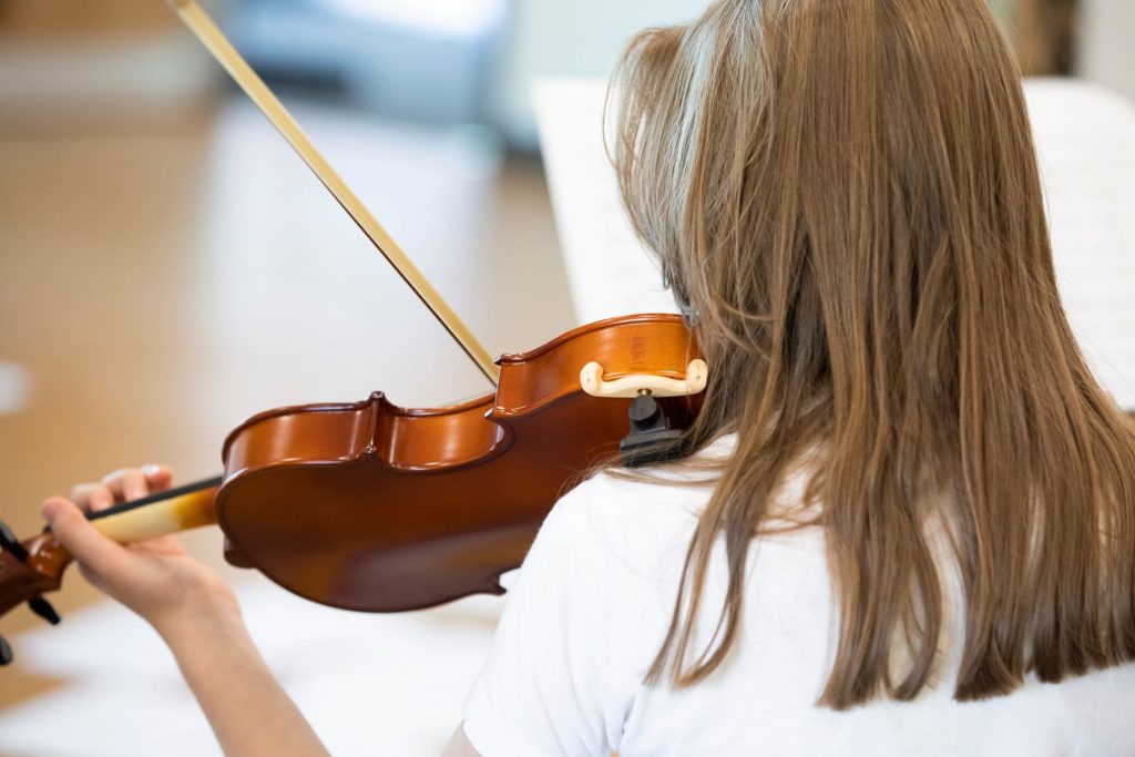 Violin.Viola A person with shoulder-length light brown hair, wearing a white shirt, plays a violin. The photo is taken from behind, showing the violin and bow in use. Sheet music is visible in the background.
