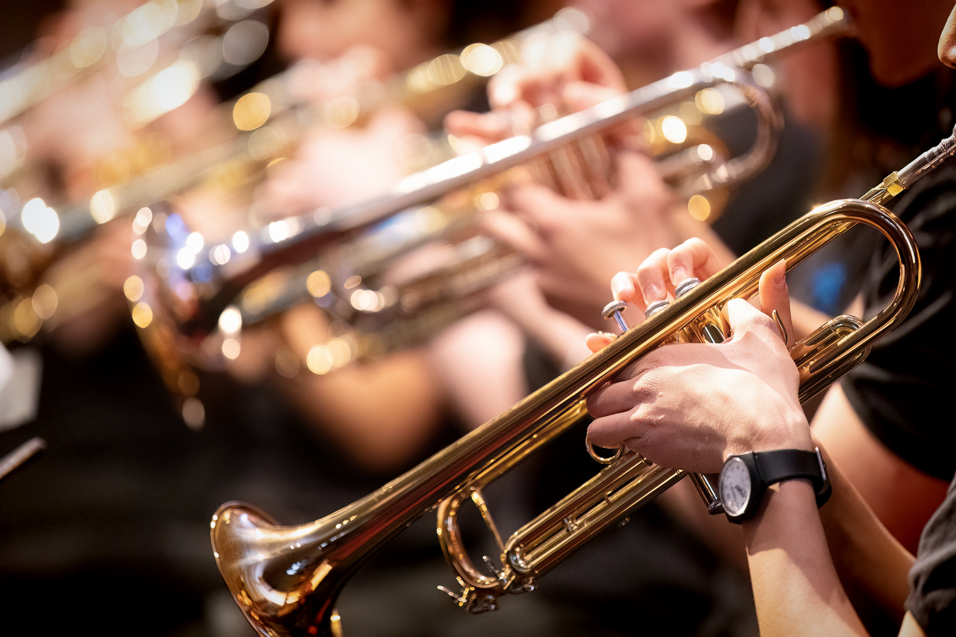 A close-up of several musicians playing trumpets in a band, with hands and shiny brass instruments in focus and people blurred in the background.