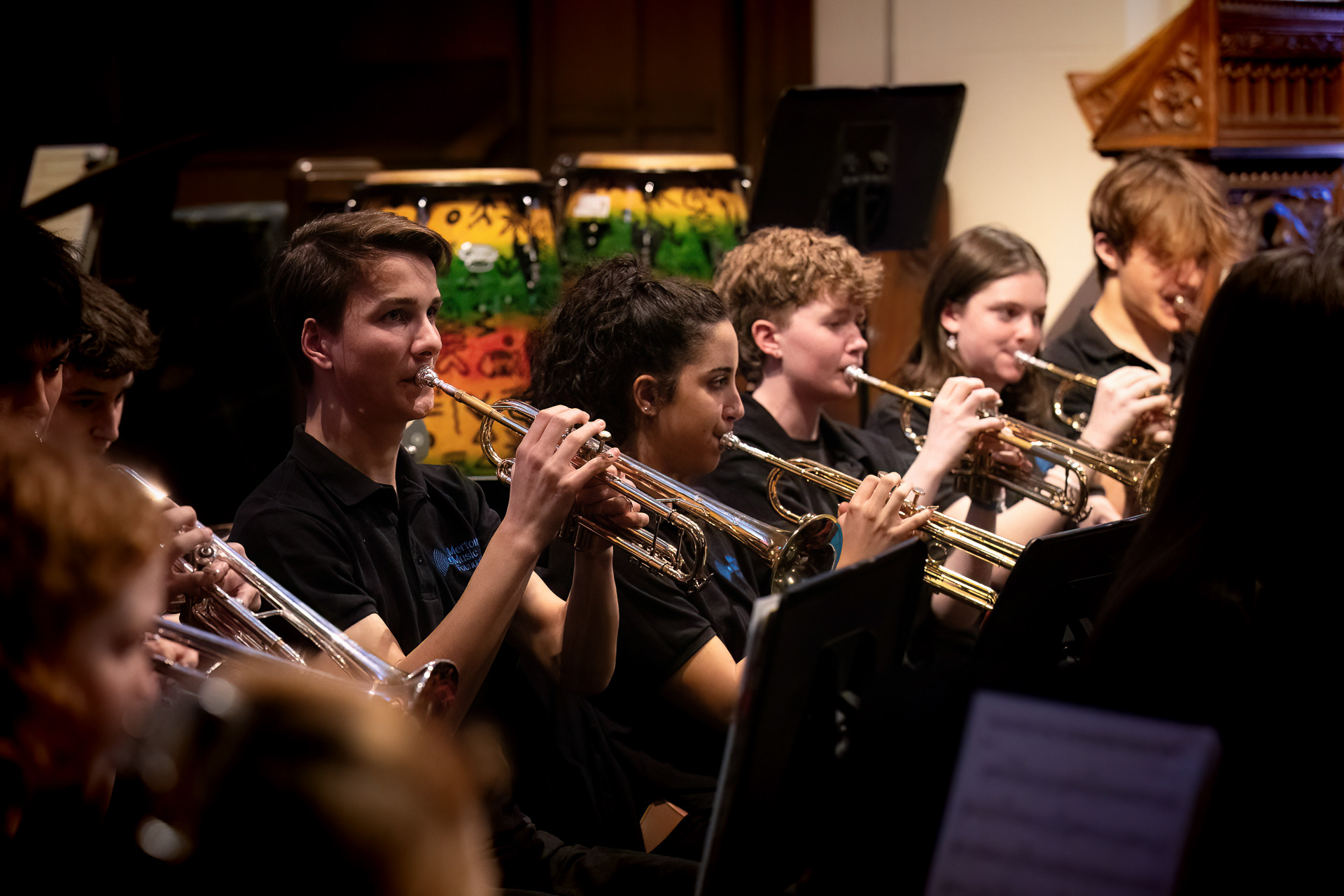 A group of young musicians from the Merton Music Foundation in black shirts play trumpets in an indoor ensemble, with focused expressions and colorful drums visible in the background.