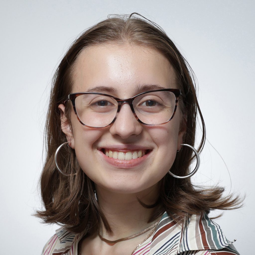 Astrid.sq_.web_-scaled A young person with shoulder-length brown hair, glasses, and hoop earrings smiles at the camera. Wearing a striped shirt and a necklace, they exude confidence—a proud member of the Youth Council—against a plain light background.