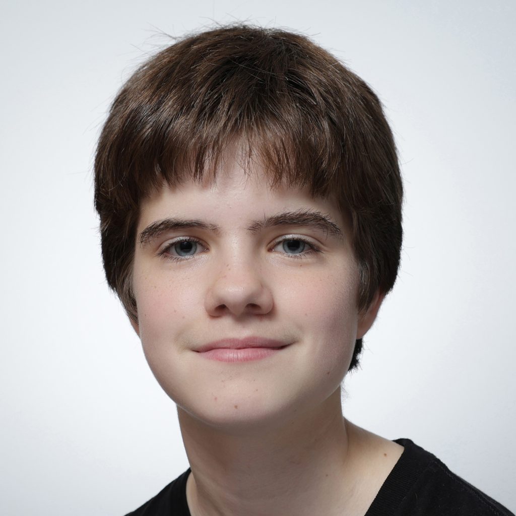 Caitlin.sq_.web_-scaled A young person with short brown hair and light skin smiles softly, looking at the camera against a plain white background. They are wearing a black shirt, representing the Youth Council with a calm and confident demeanor.