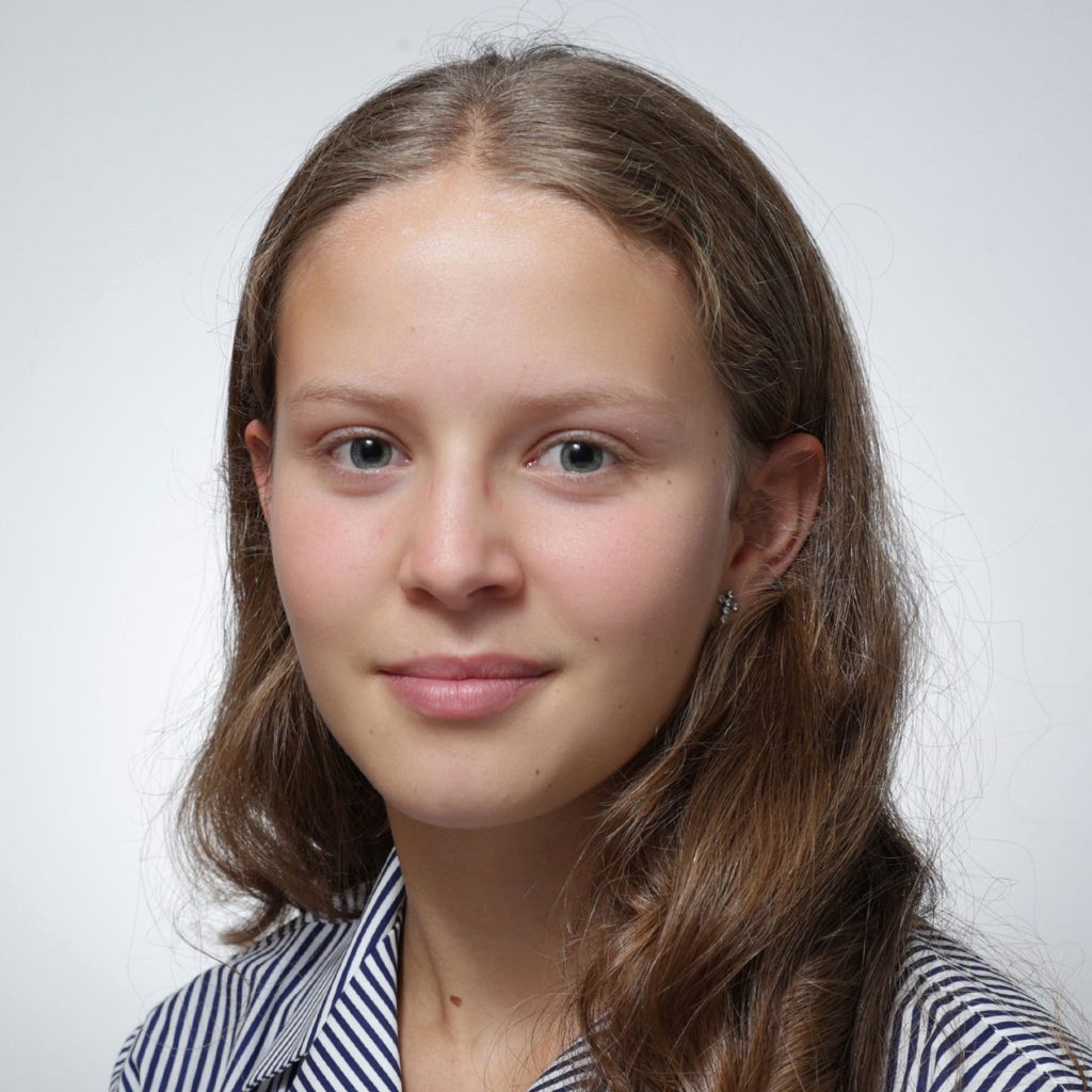 Carolina.sq_.web_-scaled A young girl with long brown hair and light skin, a member of the Youth Council, looks at the camera with a neutral expression. She is wearing a striped shirt and small stud earrings, against a plain white background.