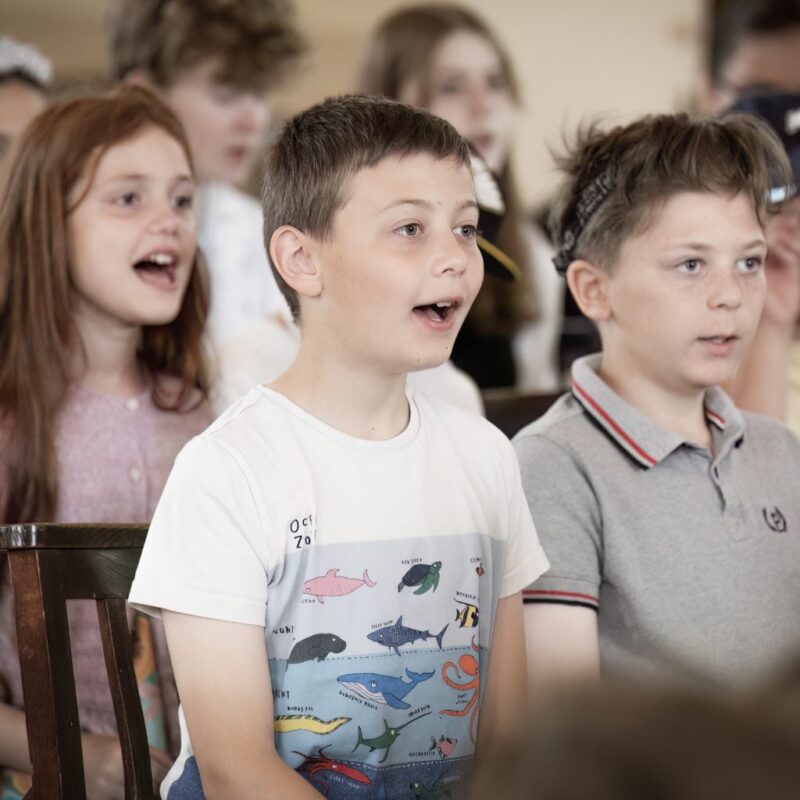 A group of children sit closely together, singing or speaking with animated expressions. The scene appears lively and engaging.