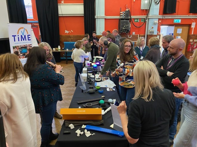 A group of people gather around tables displaying various TiME tech gadgets and devices at an indoor event. Some are talking and examining items, while banners and informational materials are visible in the background.