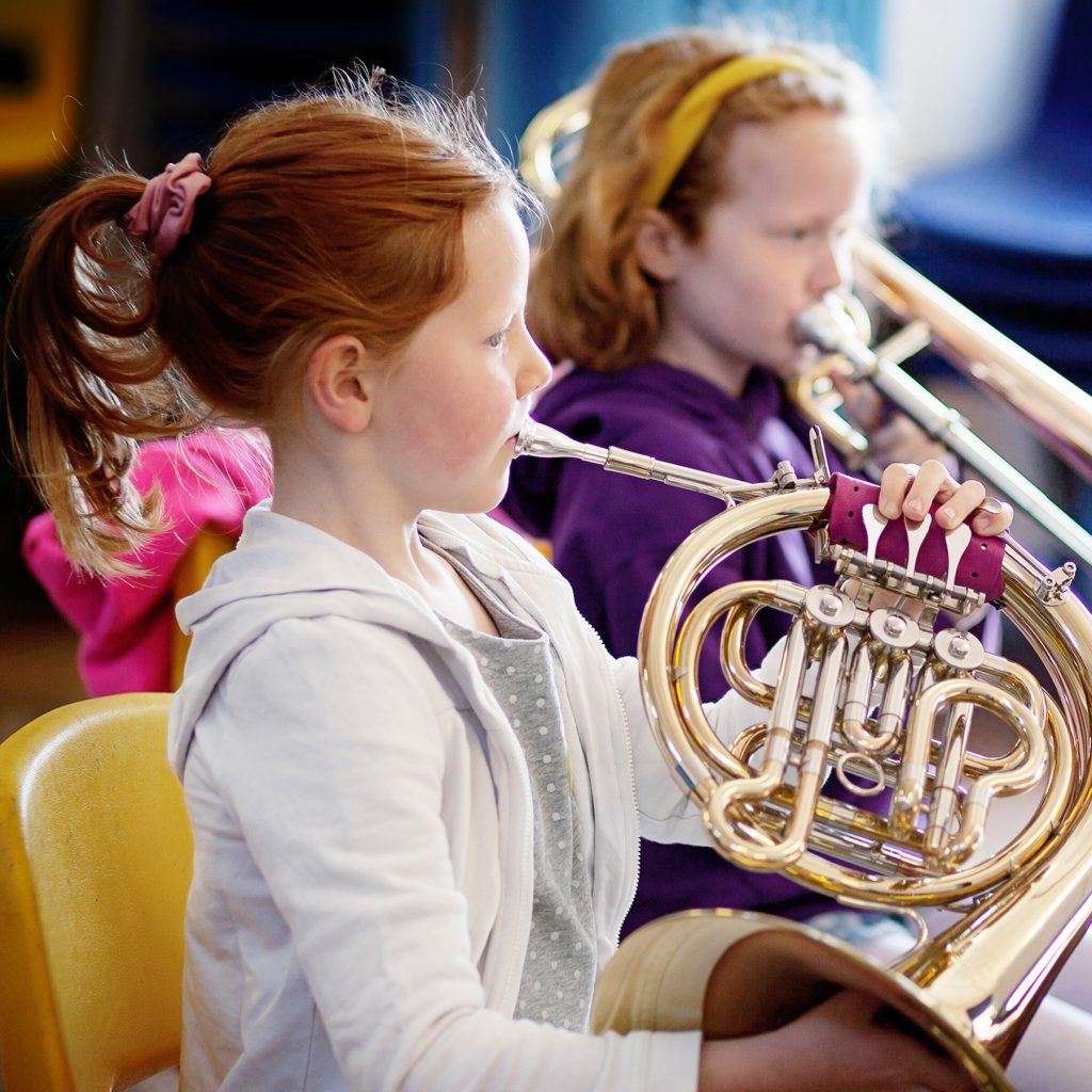 Two young girls with light skin are sitting and playing brass instruments, one playing a French horn in the foreground and the other playing a trombone in the background. Both appear focused on their music.