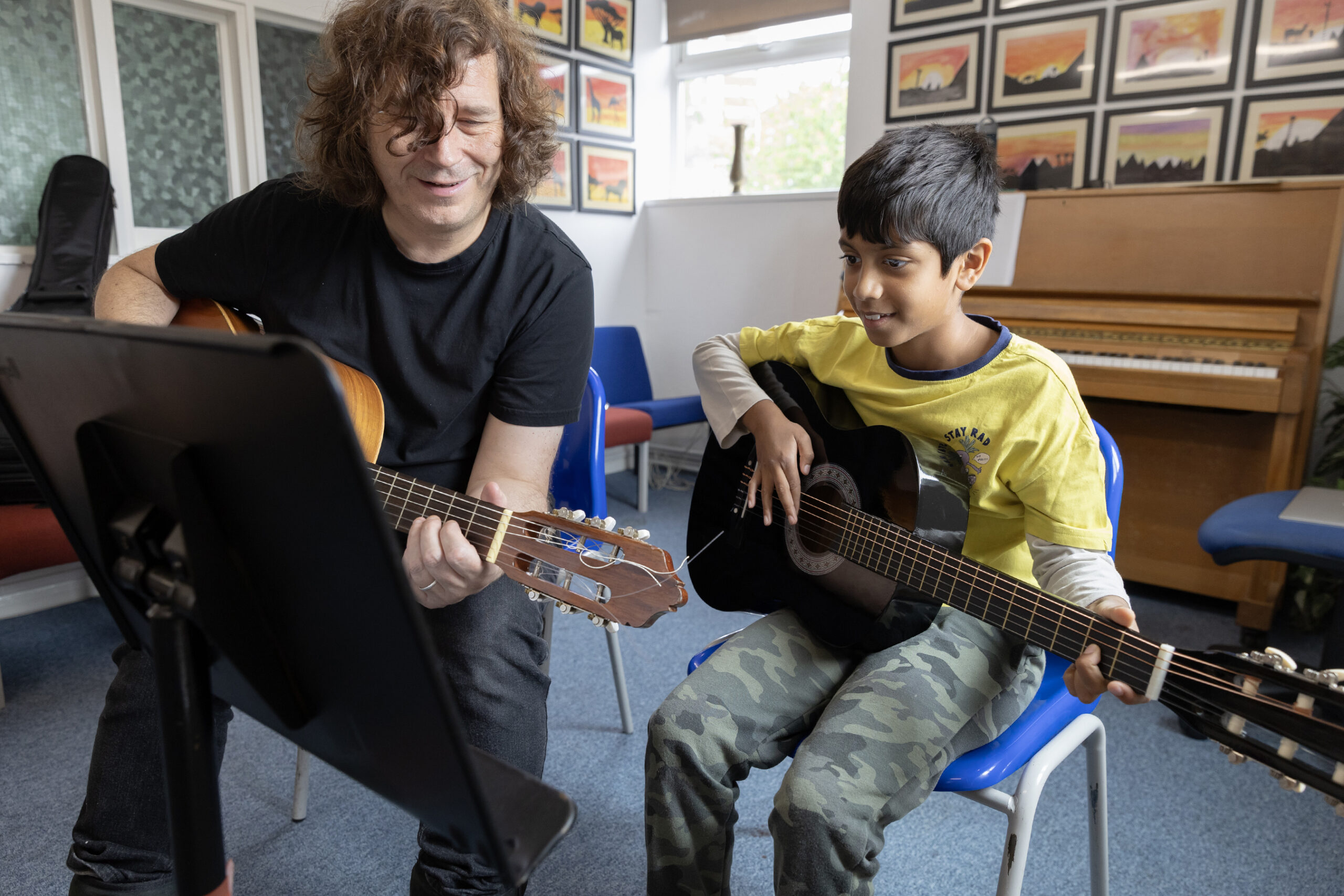 An adult and a child sit on chairs playing guitars together in a classroom decorated with colorful artwork and a piano in the background. Both appear focused and happy as they enjoy music with the Merton Music Foundation.