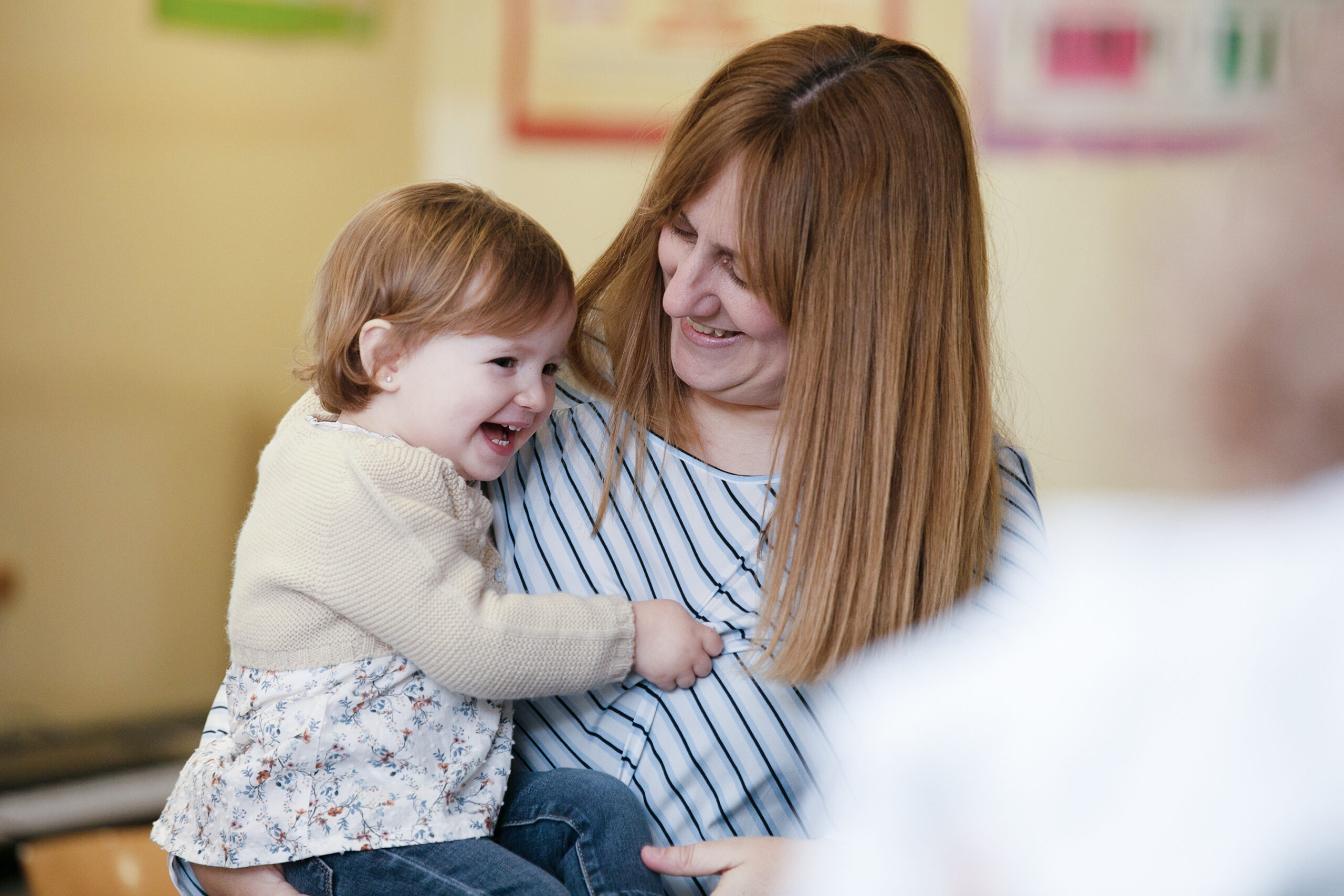 A woman with long brown hair holds a laughing toddler in her arms. Both are smiling and looking at each other in a warmly lit indoor setting, radiating joy and affection—capturing the spirit of community at Merton Music Foundation.