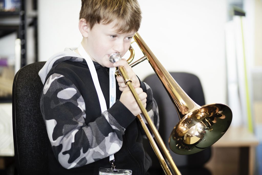 A young boy wearing a black and gray hoodie plays a brass trombone while seated indoors. He focuses intently on the instrument, showcasing his skills learned at local Music Centres.