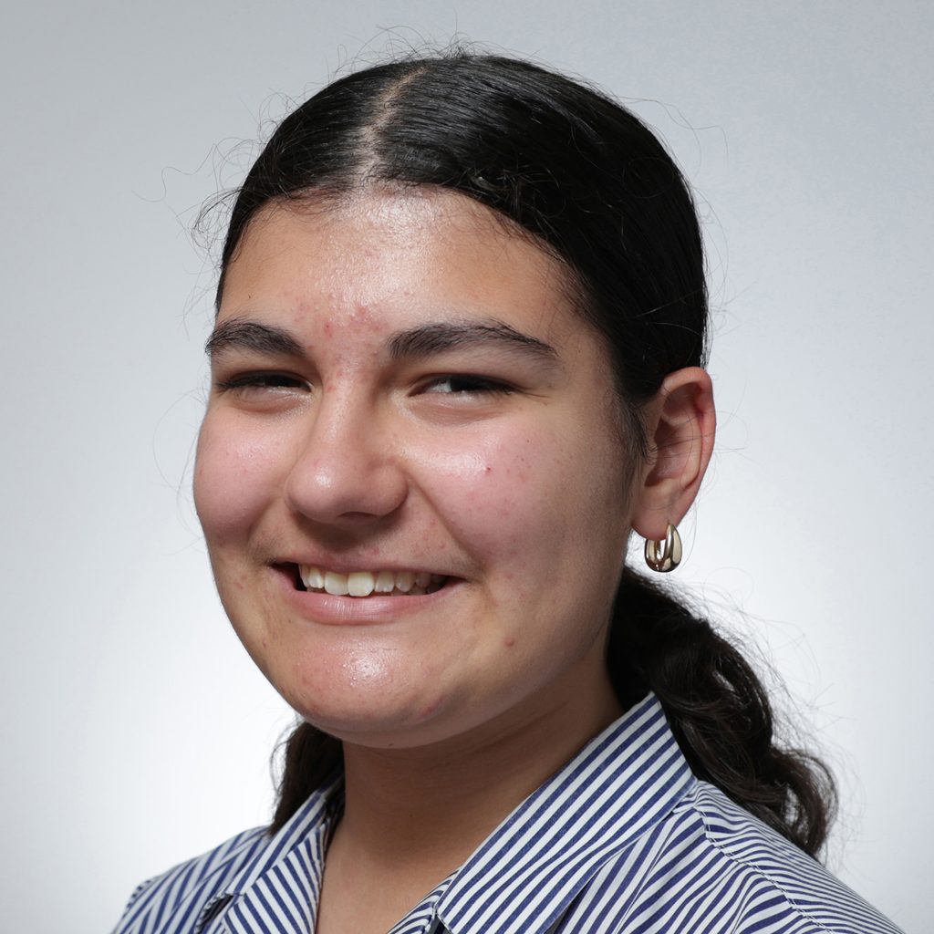 Eugenia.sq_.web_-scaled A young person with long dark hair tied back, wearing a striped collared shirt and gold hoop earrings, smiles at the camera against a plain light gray background, representing the Youth Council.