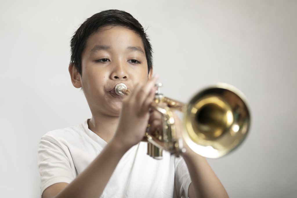 In2Music.Trumpet.June22RCP_3090.COL_-scaled A young boy in a white shirt, possibly from Merton Schools, is playing a brass trumpet. He is focused, with both hands holding the instrument against a plain, light-colored background.