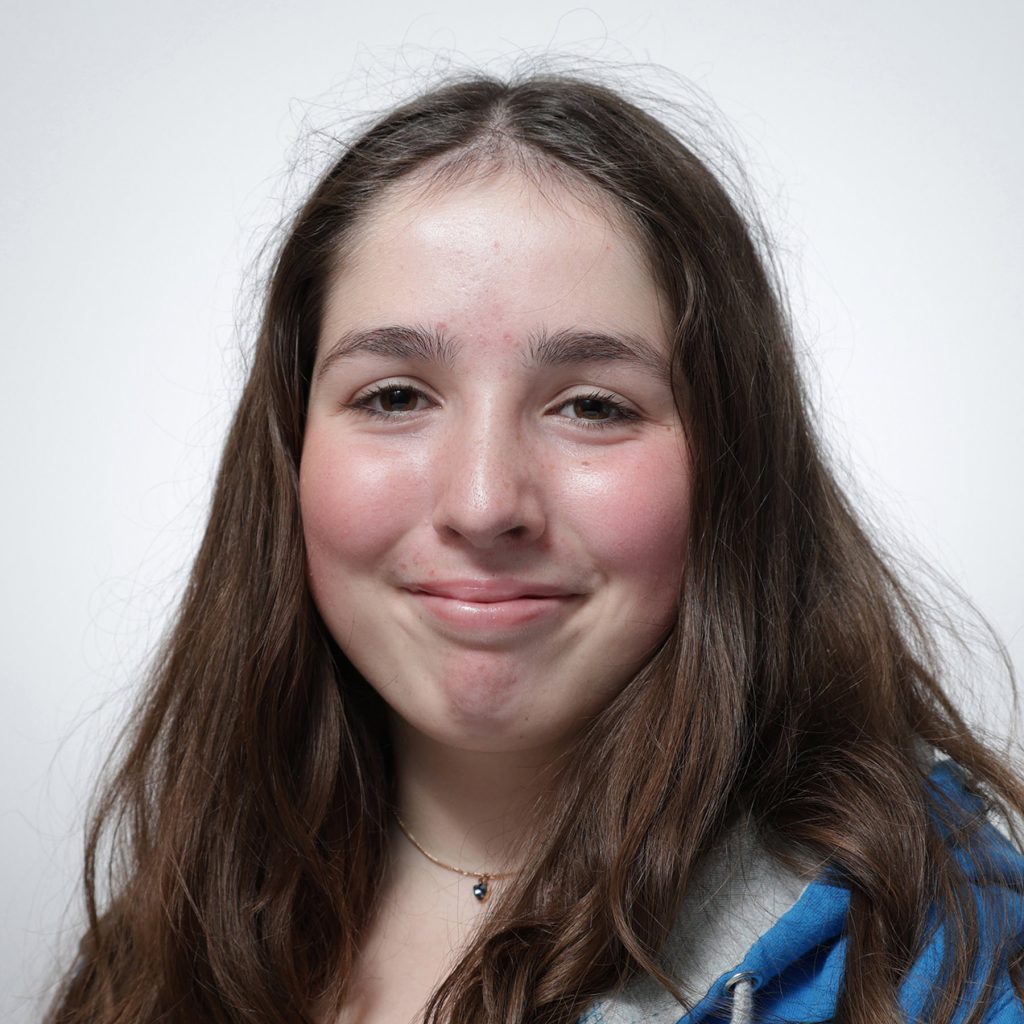 Jemima.sq_.web_-scaled A young woman with long brown hair smiles softly at the camera. She wears a blue jacket over a grey hoodie and a small necklace, representing the Youth Council. The background is plain white.
