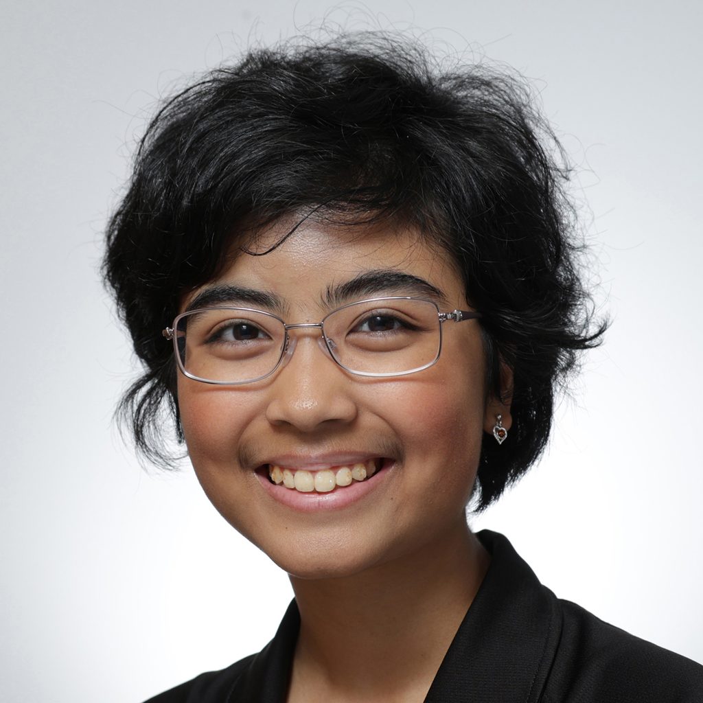Kiran.sq_.web_-scaled A young person with short, dark, wavy hair wearing glasses and a black blazer smiles at the camera against a plain light background, representing their role on the Youth Council.