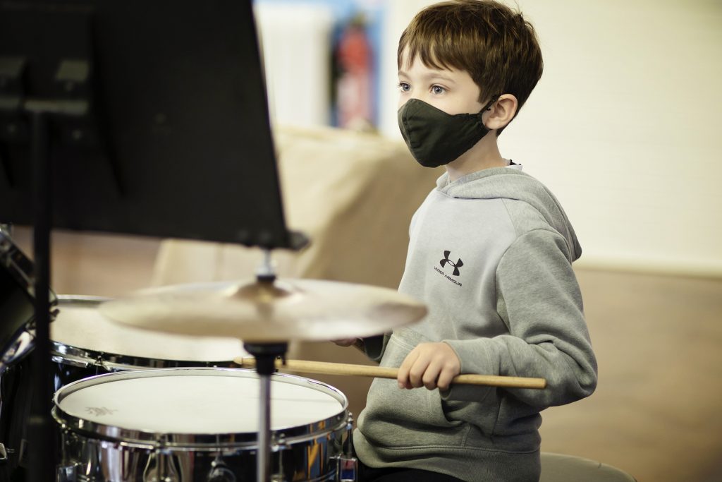 A young boy wearing a grey hoodie and a face mask plays a drum set at one of the local Music Centres, holding drumsticks and looking at sheet music on a stand in front of him.