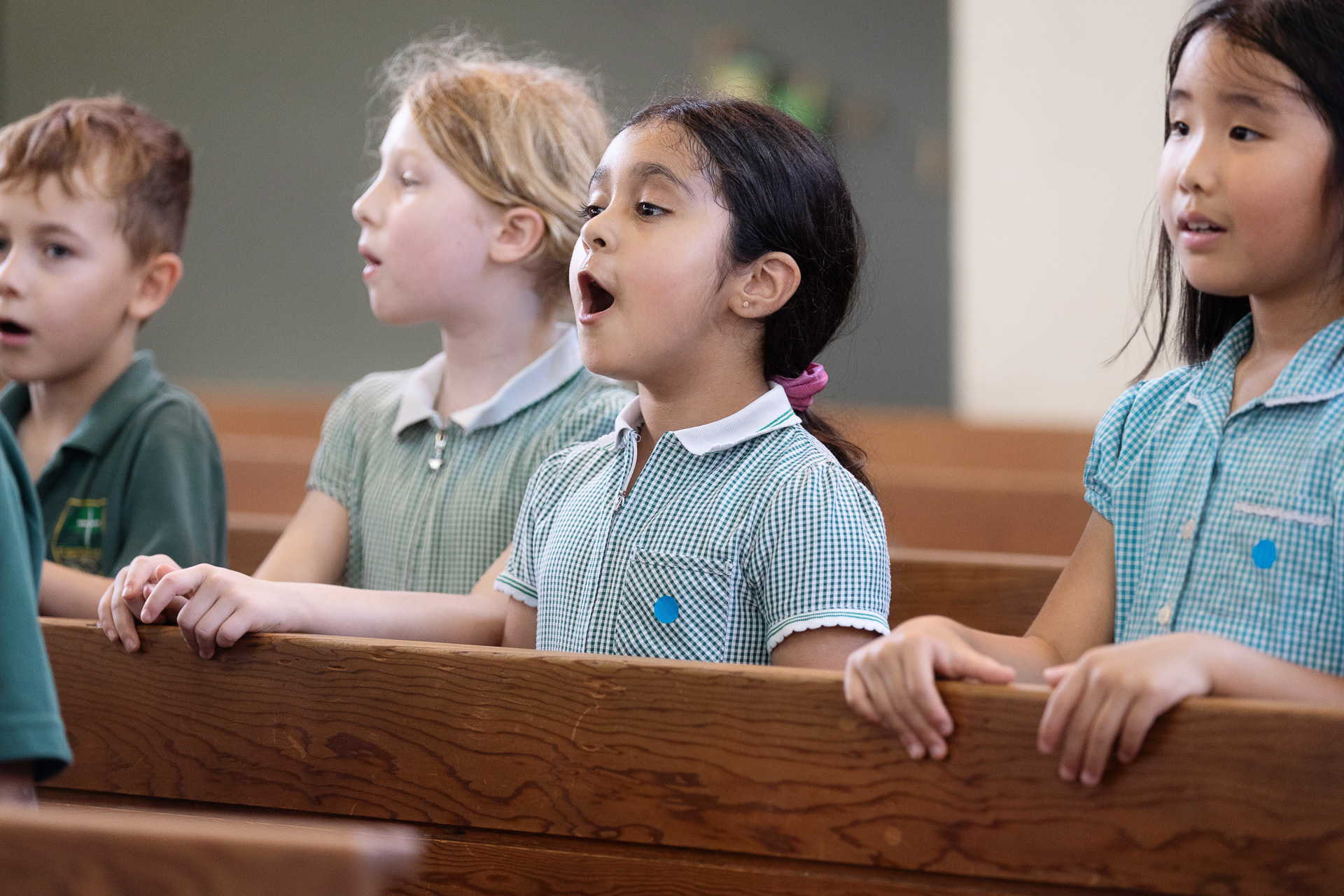 Four young children sit in wooden pews, singing together. They wear green-checkered school uniforms and appear to be in a classroom or assembly setting, with focused and expressive faces.