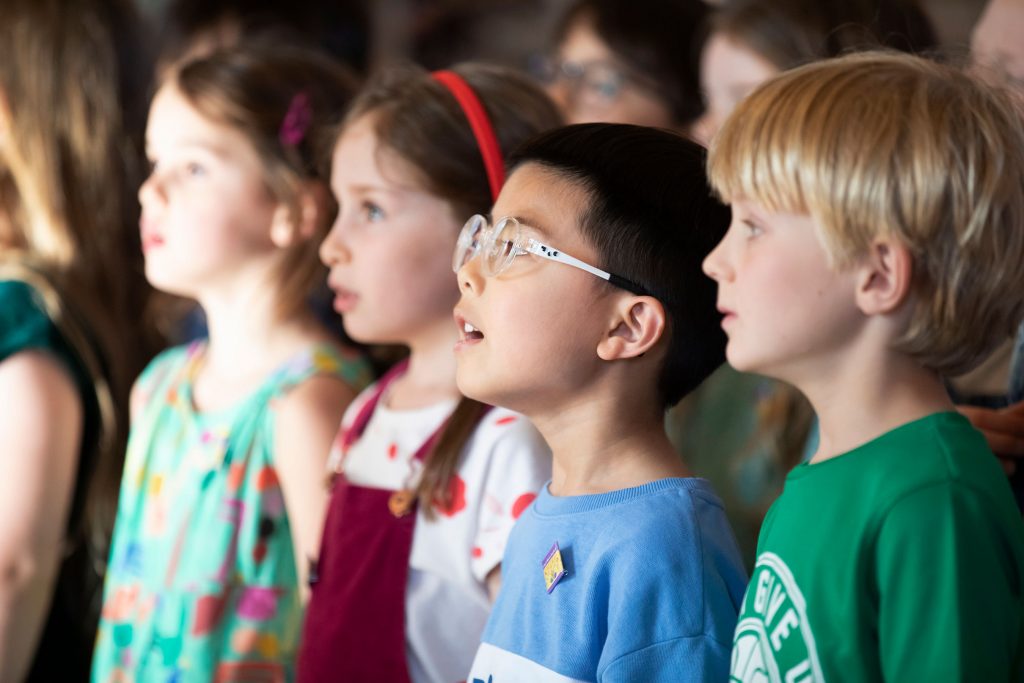 A group of young children stand closely together, looking ahead with attentive and amazed expressions, as if watching a performance or listening intently at one of the local Music Centres.