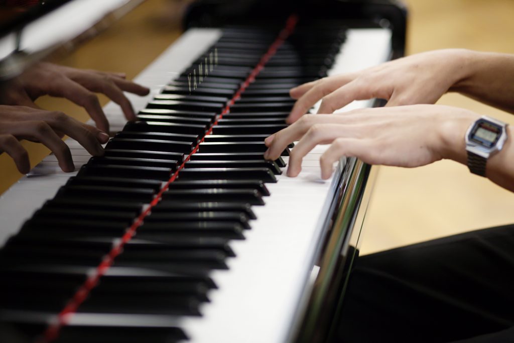 Close-up of hands playing a piano keyboard at one of the local Music Centres, with fingers pressing the keys and reflections visible on the glossy black surface. The person wears a digital watch on their left wrist.