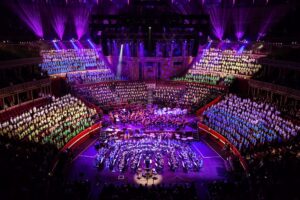 A large orchestra performs on stage as hundreds of choir members, arranged in colourful sections, fill the balconies of a grand Music Centre, all illuminated by dramatic purple lighting.