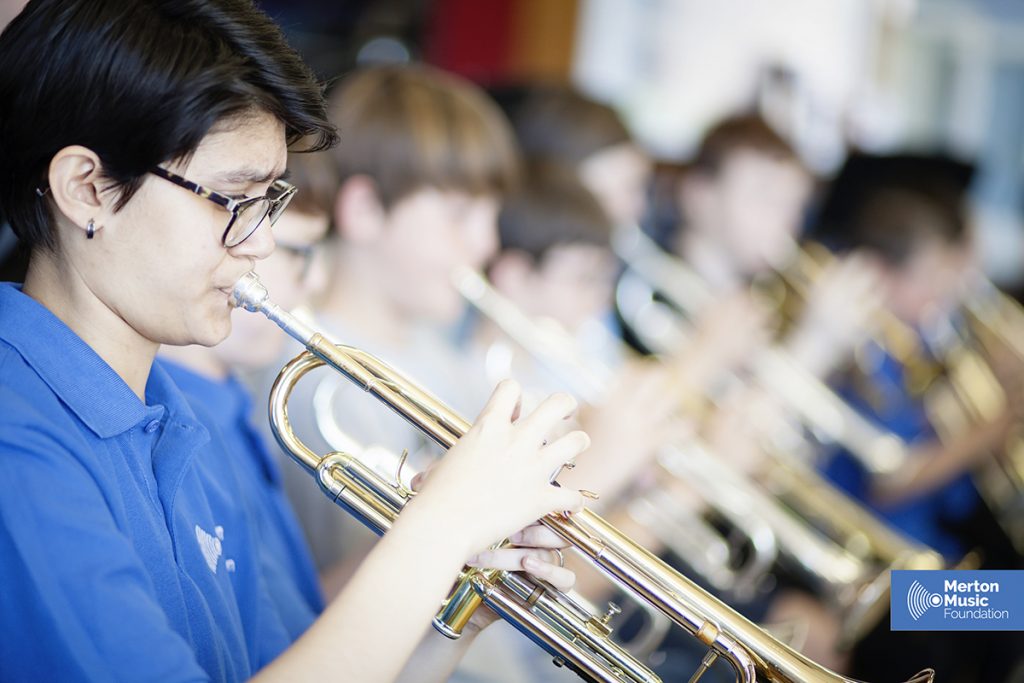 A group of young musicians in blue shirts play trumpets in a brightly lit room, representing one of the Music Centres. The focus is on one player in the foreground, with the Merton Music Foundation logo visible.
