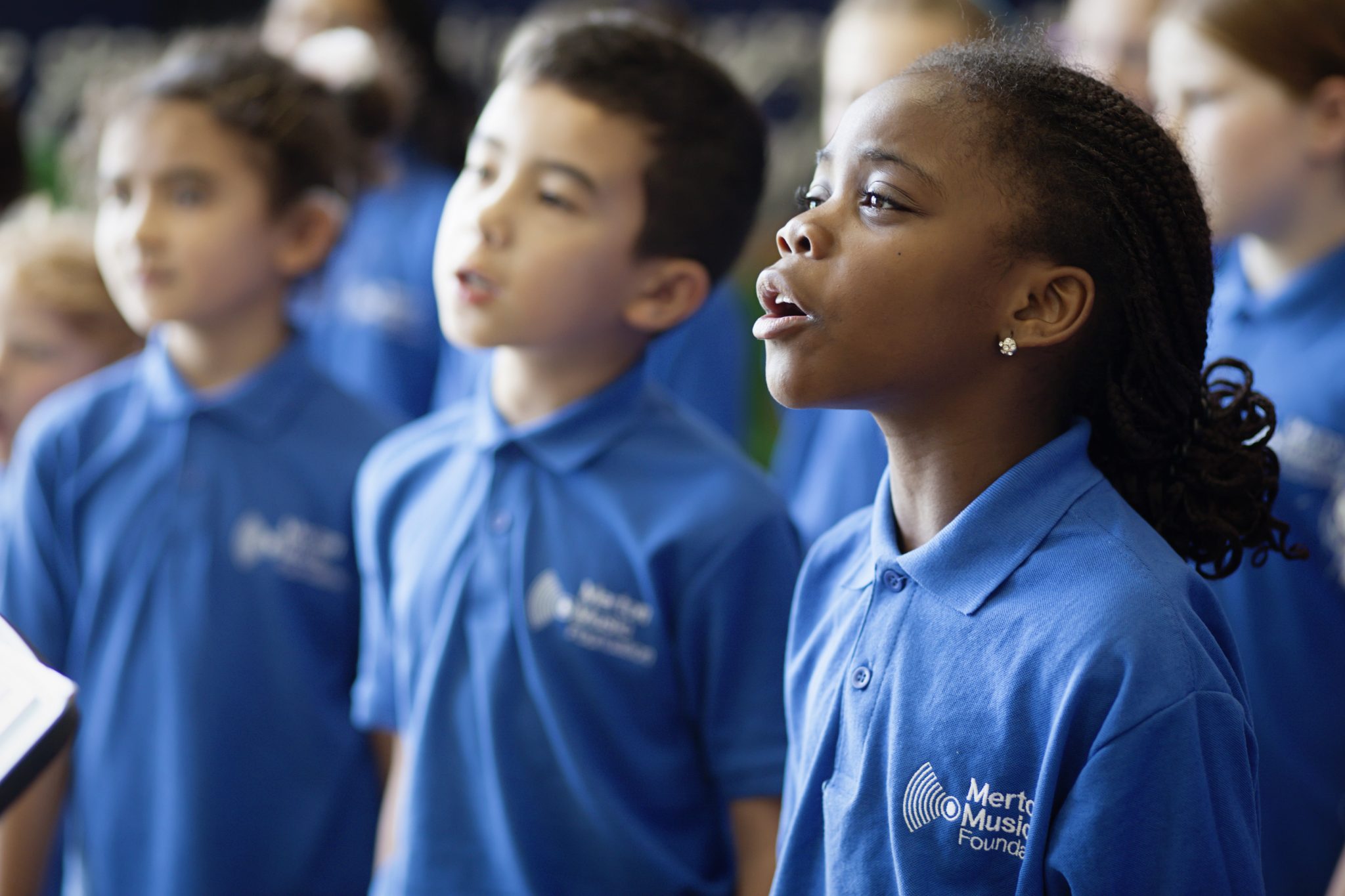 A group of children wearing blue Merton Music Foundation polo shirts sing together, focusing intently. The scene radiates inclusion, with one girl in the foreground singing passionately.