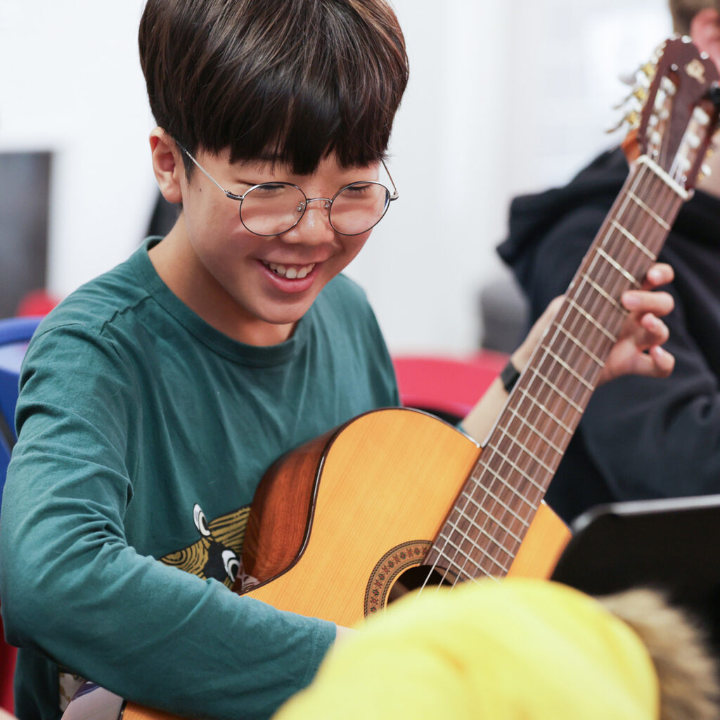 A young person with glasses smiles while playing an acoustic guitar, seated indoors in a casual setting, practicing for the Senior Guitar Ensemble.