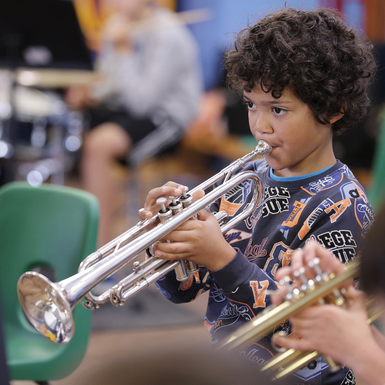 mmfmusiccentre0011web_54369298823_o.sq.JuniorConcertBand A young boy with curly hair plays a silver trumpet in a Junior Concert Band classroom, focusing intently. Other students and musical instruments are visible in the blurred background.