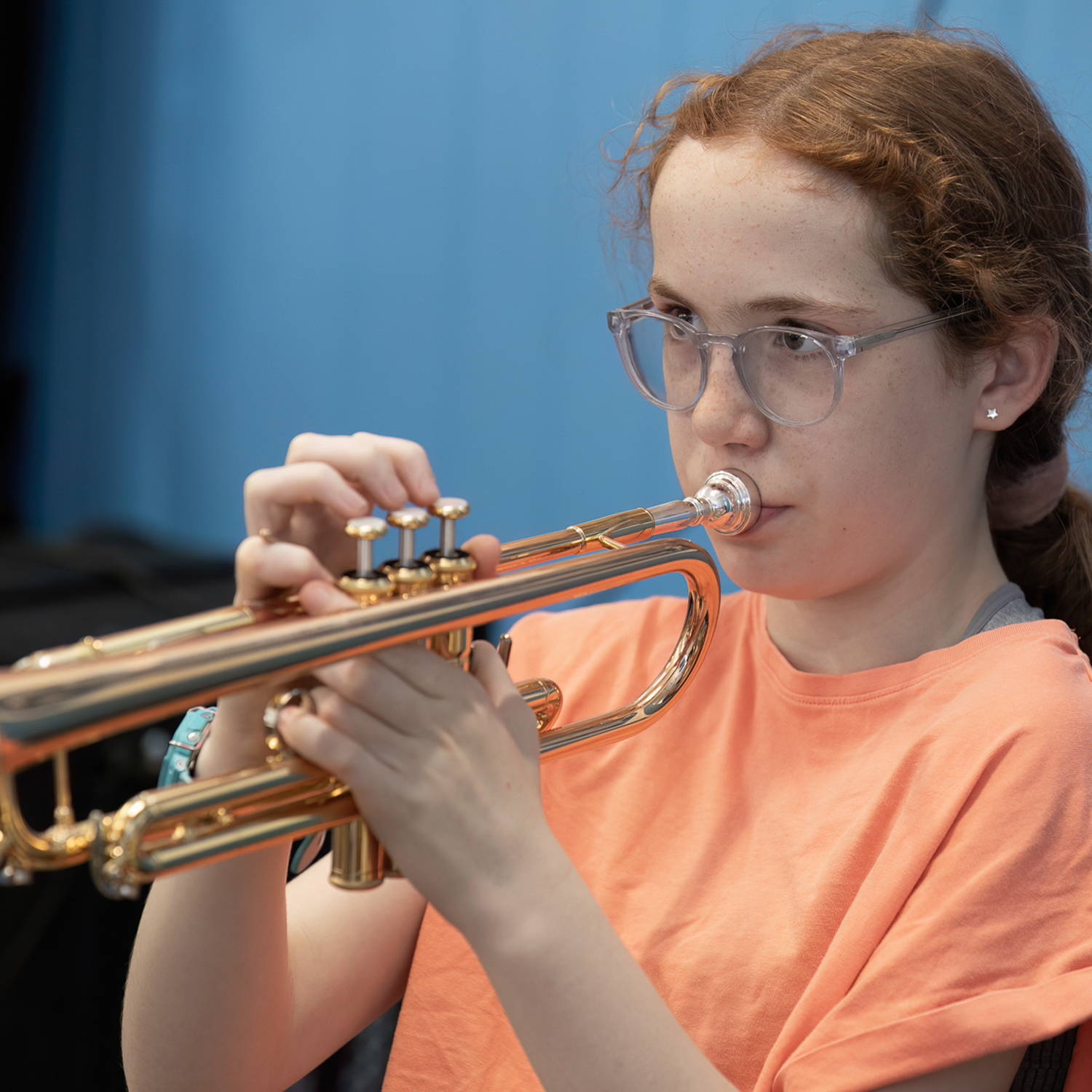 rcp_0805web_54369460945_o.sq.ICB A young person with glasses and red hair, wearing an orange shirt, is playing a brass trumpet against a blue background, perfect for an Intermediate Concert Band performance.