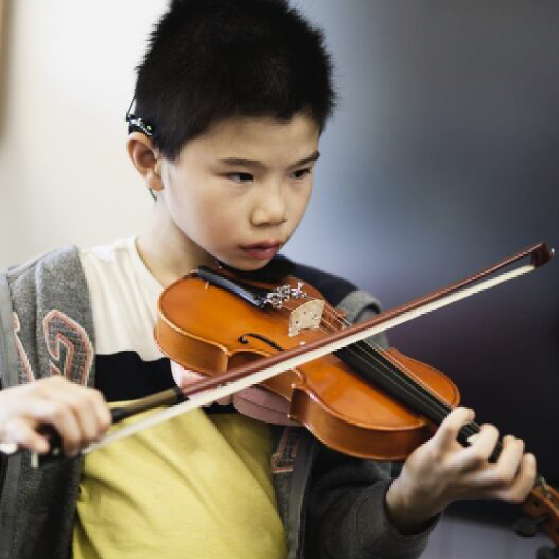A young boy with short black hair, part of the String Busters, plays the violin with focus on the strings. He wears a yellow shirt, a gray jacket, and has a hearing aid behind his ear.
