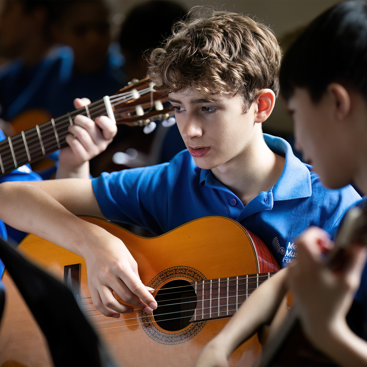 A teenage boy in a blue shirt plays an acoustic guitar in an Intermediate Guitar Ensemble, focusing intently. Other students with guitars are nearby, suggesting a music class or practice session.