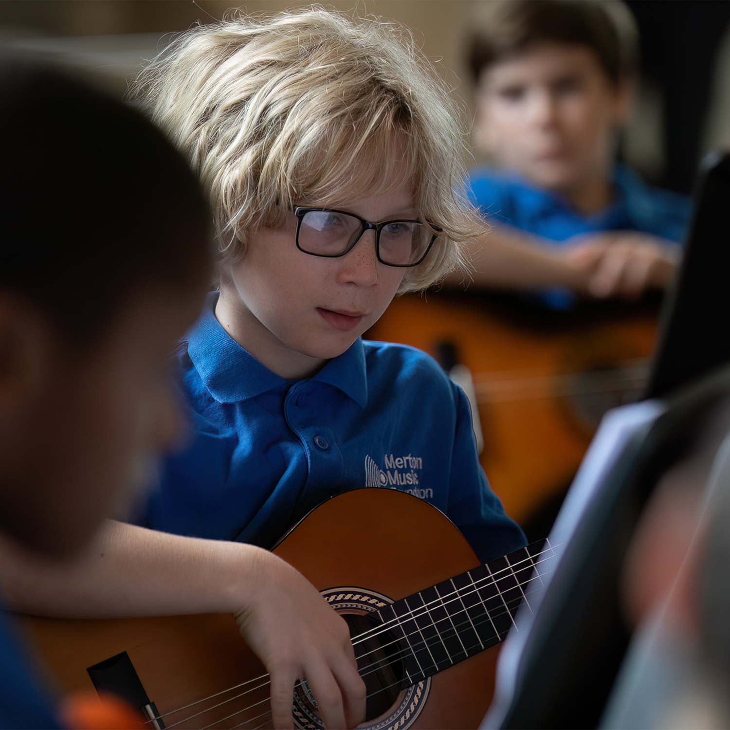 summerconcert23110web_54369460535_o.sq.JuniorGuitars A young boy with blond hair and glasses, wearing a blue polo shirt, plays an acoustic guitar and reads music as part of the Junior Guitar Ensemble. Two other children with guitars are blurred in the background.