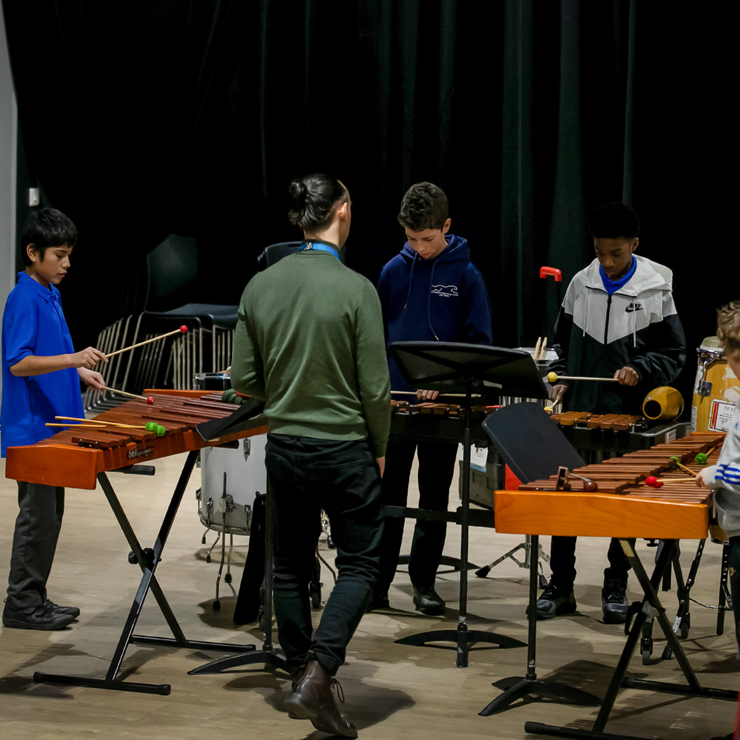 Four boys stand around xylophones and marimbas in a dimly lit music room, performing as the Merton Mallets. An adult observes while music stands and percussion instruments are visible in the background.
