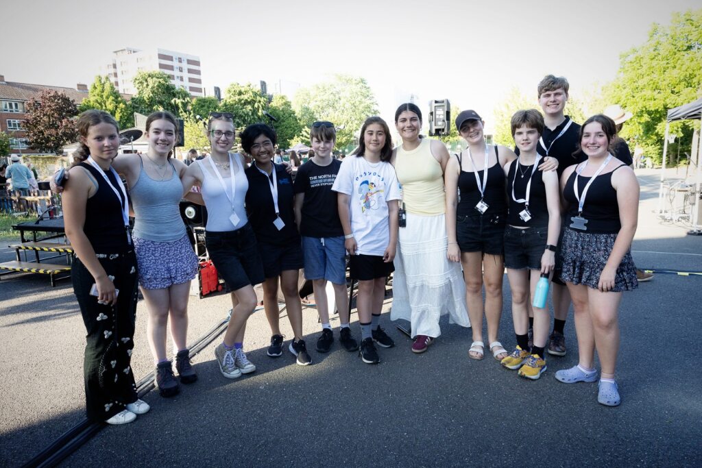 Youth Council A group of eleven smiling teenagers from the Youth Council pose together outdoors on a sunny day. They wear casual summer clothes and name tags, standing on a paved area with greenery and buildings in the background.