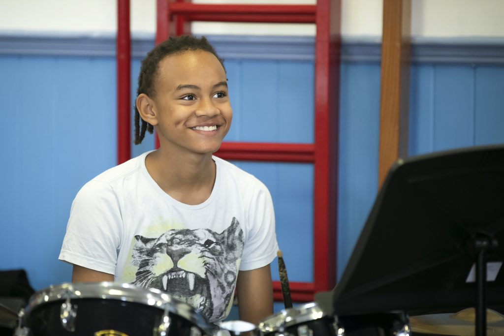 A smiling child with short braided hair and a white tiger-print t-shirt sits at a drum set in South Wimbledon, looking towards a music stand, with a blue wall and red bars in the background.