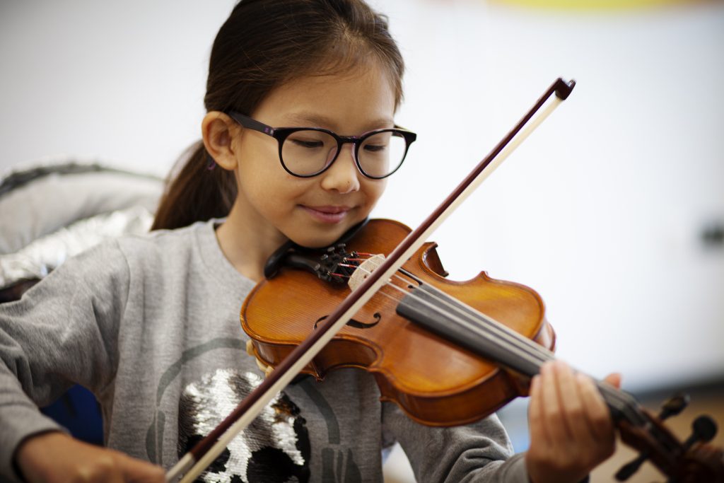 Wimbledon College 2 A young girl with glasses and a gray sweatshirt, possibly from Wimbledon College, plays the violin, smiling and focusing on her instrument against a blurred background.
