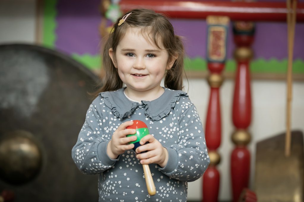Wimbledon College A young girl with brown hair and a gray polka dot sweater smiles while holding a red and green maraca in a colorful classroom setting at Wimbledon College.