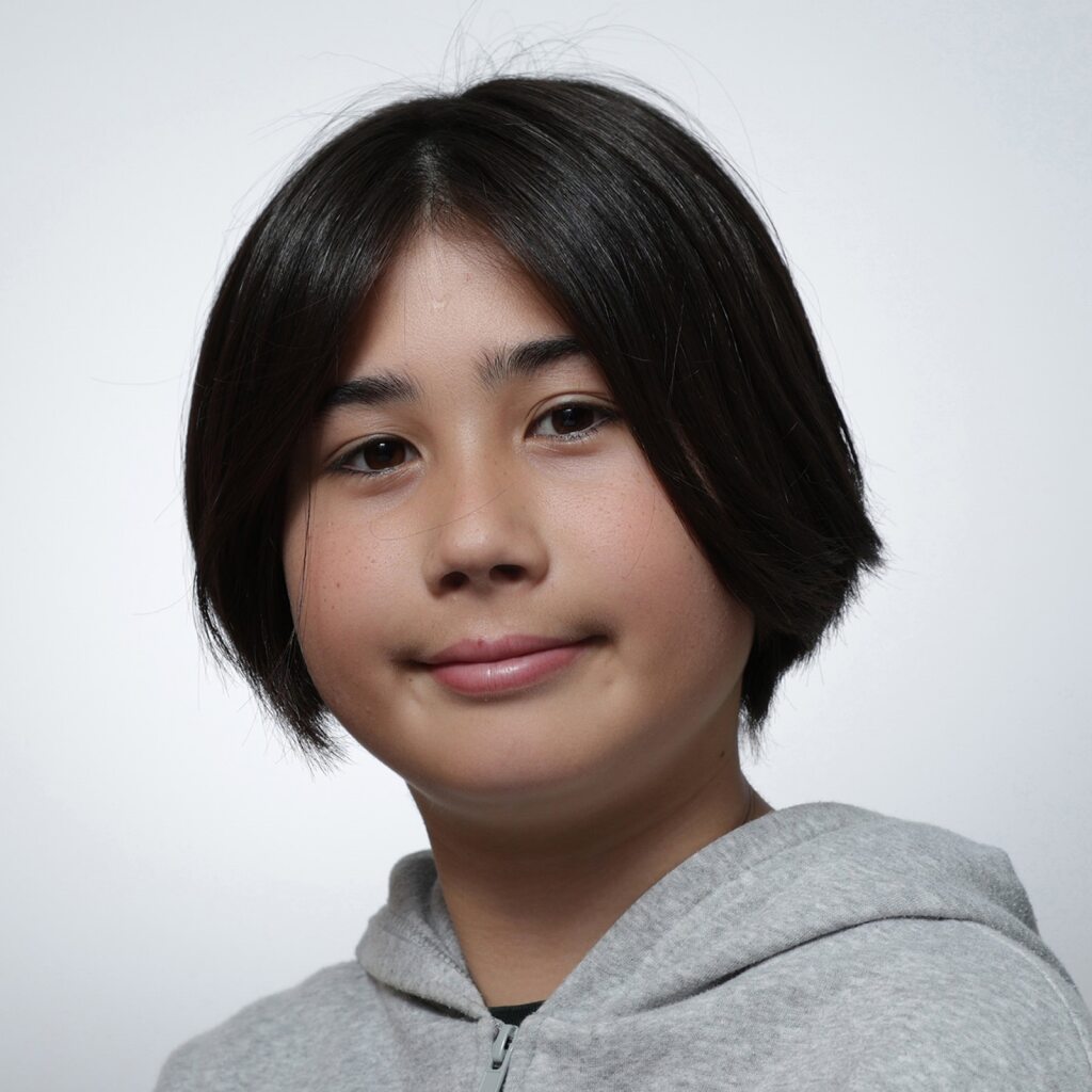 Toki A young person with straight, dark hair in a short, layered style smiles gently at the camera. Wearing a light gray hoodie, they represent the Youth Council as they pose against a plain white background.