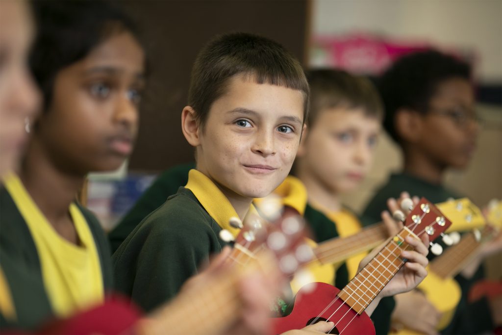 I A group of children in school uniforms play small red ukuleles in a classroom. One boy in the center looks directly at the camera and smiles, while other students focus on their instruments during a CPD music activity.