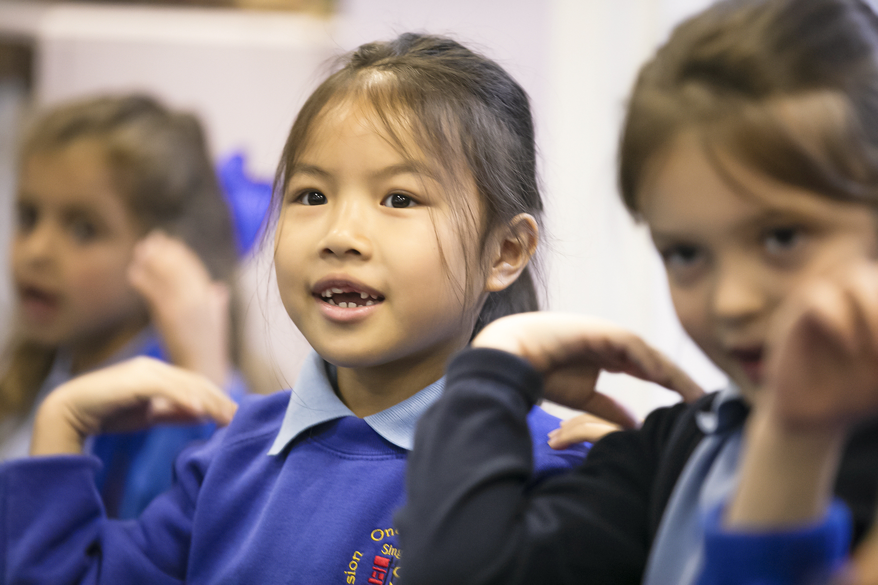 In2Music.GM_.Singlegate.02.COL_.sm_ Three young girls in blue school uniforms raise their hands to their shoulders, engaging in a CPD-inspired classroom activity. The girl in the center looks forward, smiling slightly, while the softly blurred background highlights their focus.