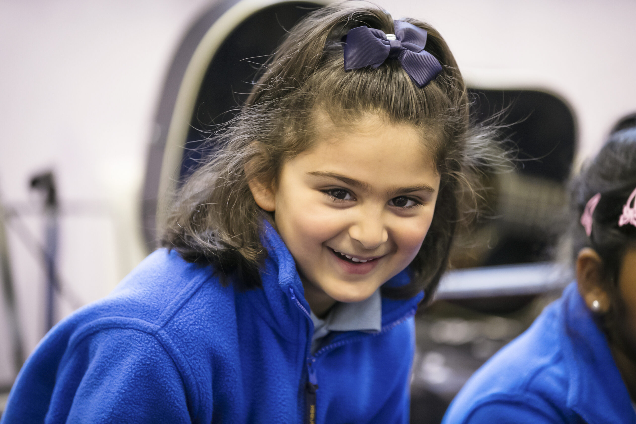 In2Music.GM_.Singlegate.03.COL_.sm_ A young girl with dark hair in a blue fleece jacket smiles at the camera, proudly wearing a blue bow in her hair at the CPD event. Another child is partially visible beside her. The background is blurred.