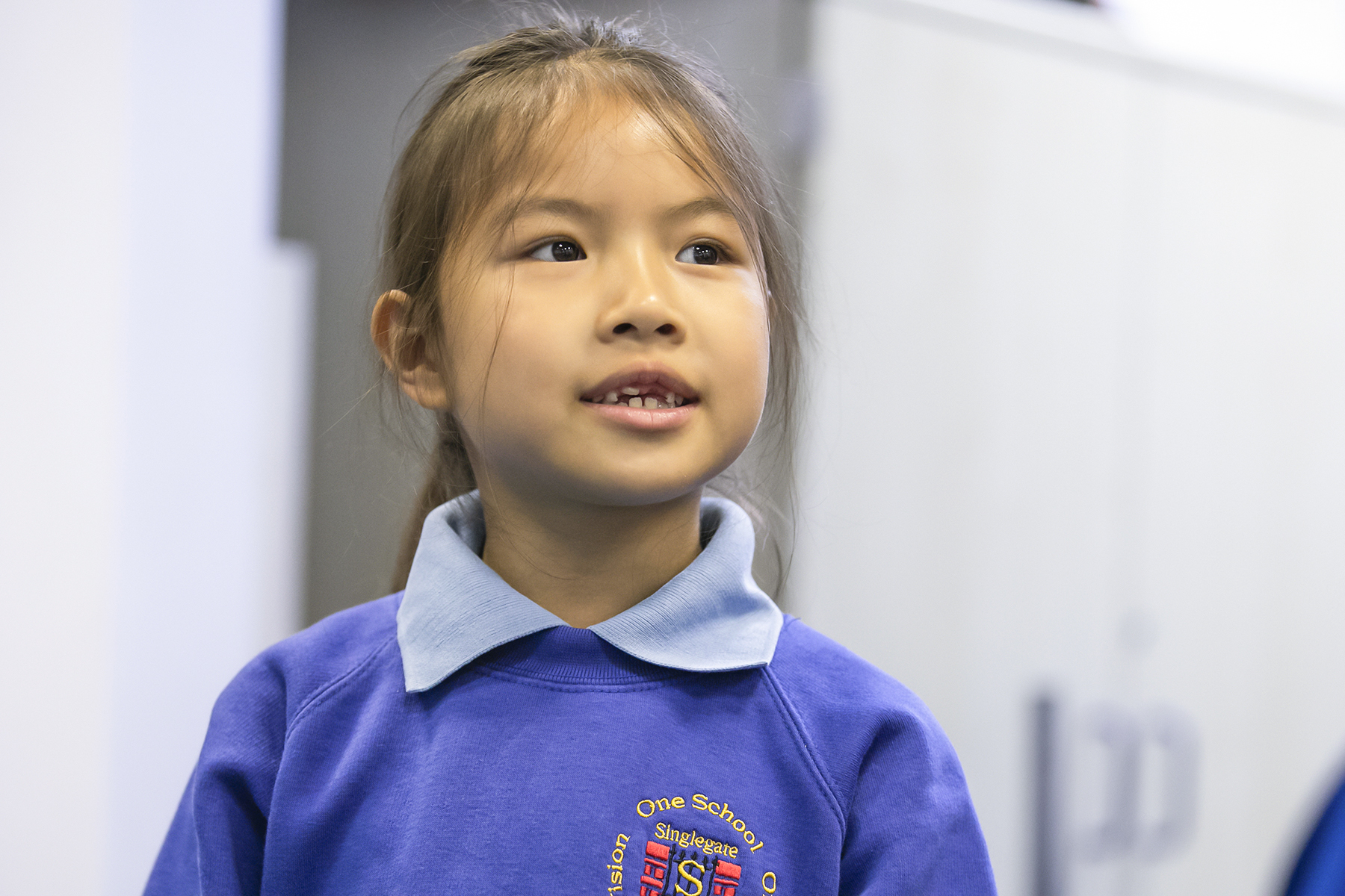 In2Music.GM_.Singlegate.05.COL_.sm_ A young girl with long brown hair, wearing a blue school sweatshirt and a light blue collar, looks slightly upward and smiles in a CPD-supported school setting.