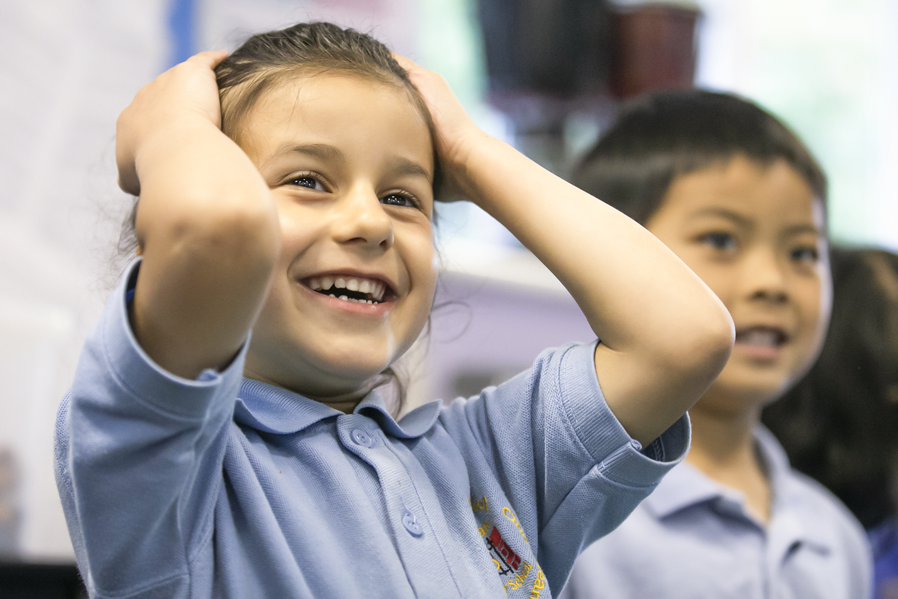 In2Music.GM_.Singlegate.07.COL_.sm_ Two young children in light blue school uniforms stand indoors. The girl in front smiles brightly with her hands on her head, while the boy behind her looks on, slightly smiling—a cheerful moment during a CPD activity. The background is blurred.