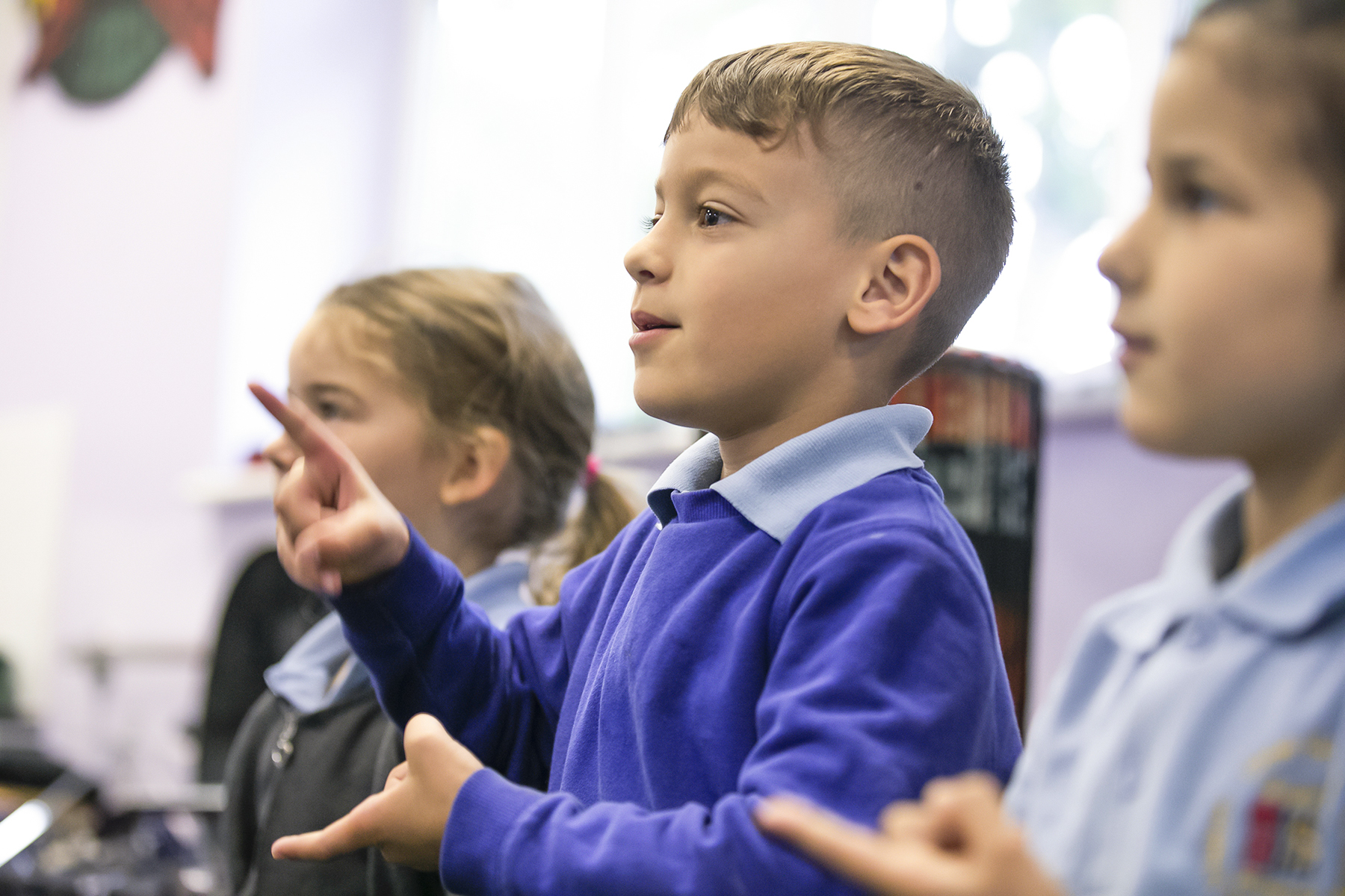In2Music.GM_.Singlegate.14.COL_.sm_ Three young children in blue uniforms are standing indoors, using their hands to sign, possibly in a classroom or CPD setting. The focus is on the boy in the center, who is actively gesturing.